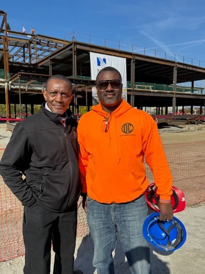 Two men on a construction site, one in an orange safety hoodie holding a blue hard hat, in front of a steel-framed building with a banner