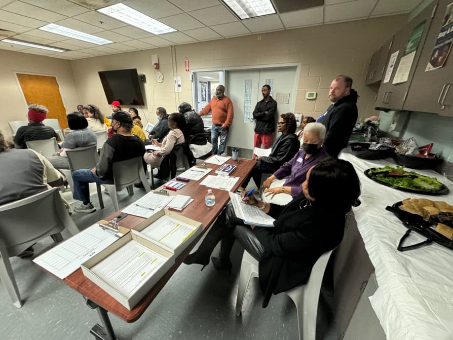 A crowded community meeting in a small room, with sign-in sheets and clipboards on a table in the foreground and a man standing to speak at the back